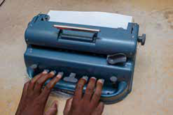 A imagem mostra mãos utilizando máquina de escrever Braille.