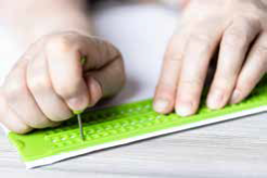 A imagem mostra mãos utilizando reglete e punção para escrita em Braille.